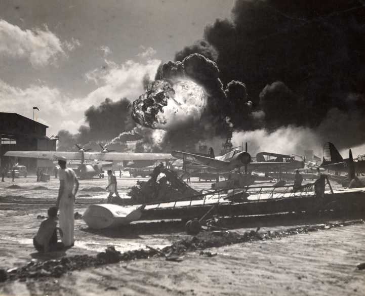 Sailors stand amid wrecked planes at the Ford Island seaplane base, watching as USS Shaw explodes in the center background